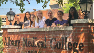 Several young women , members of Albany Medical College’s NextGen Neuroscience Virtual Summer Program, standing outside behind a brick wall at Albany Medical College.