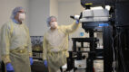 Two men wearing lab gowns, hair nets, masks, and gloves look at a large microscope