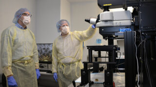 Two men wearing lab gowns, hair nets, masks, and gloves look at a large microscope