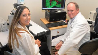 Scientists Whitney Longmate, PhD, and Michael DiPersio, PhD, wearing white coats and sitting near a computer with a green and red image