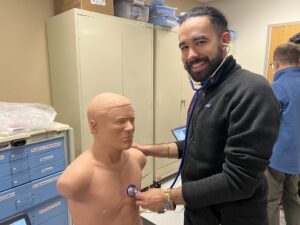 A pediatric resident, smiling, practices listening to a heart on a mannequin 
