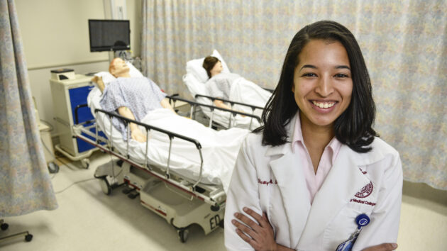 A medical professional standing in a specialized clinical training room outfitted with hospital beds and equipment.