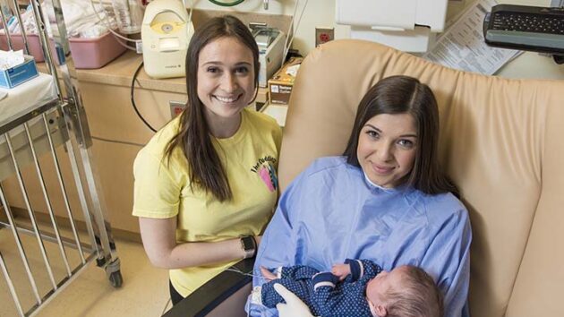 An Albany Medical Center staff member in a yellow shirt stands to the left of a woman in a hospital bed holding a newborn baby.