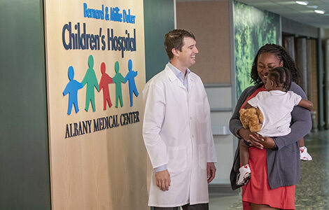 A Department of Pediatrics doctors stands in the children's hospital hallway and speaks to a mom holding a young boy.