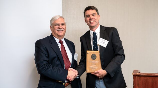 A man presents an award to another man. The two smile and pose for a picture.