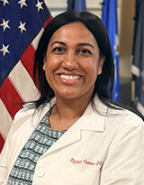 Portrait of endocrinologist Dr. Elizabeth Oommen, wearing a white coat with the American flag in the background