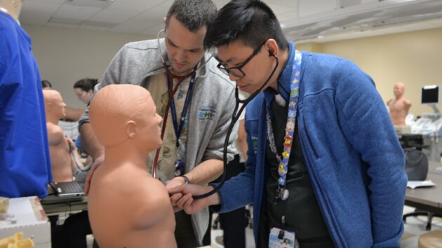 Two pediatric residents practice listening to a patient's heart using a mannequin.